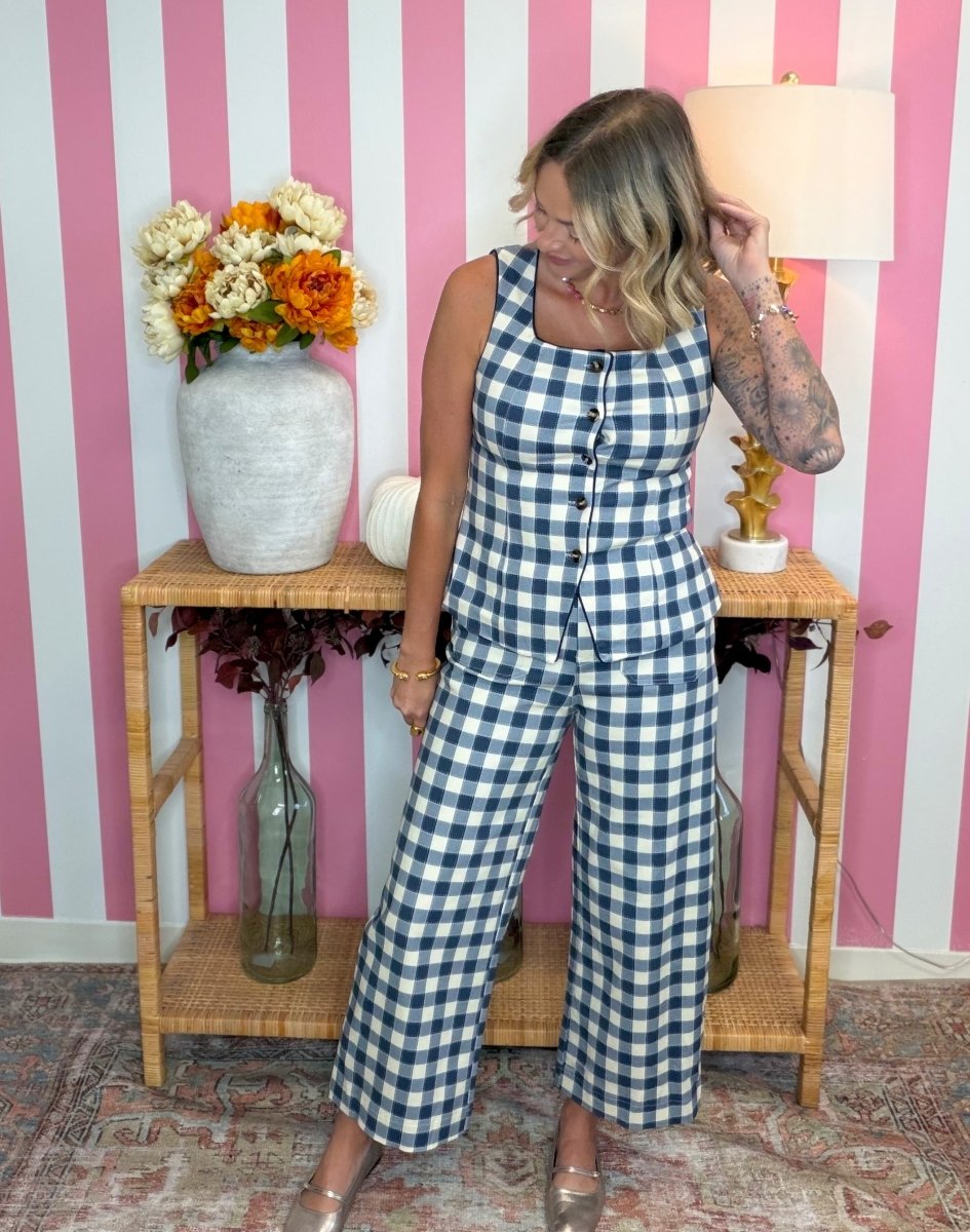 A woman models a blue and white gingham check two-piece jumpsuit with a button-front bodysuit top and wide-leg pants, standing in front of a pink and white striped wall.