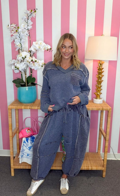 A woman modeling a blue denim hooded jumpsuit in front of a pink and white striped wall, with a wooden side table, lamp, and potted orchid in the background.