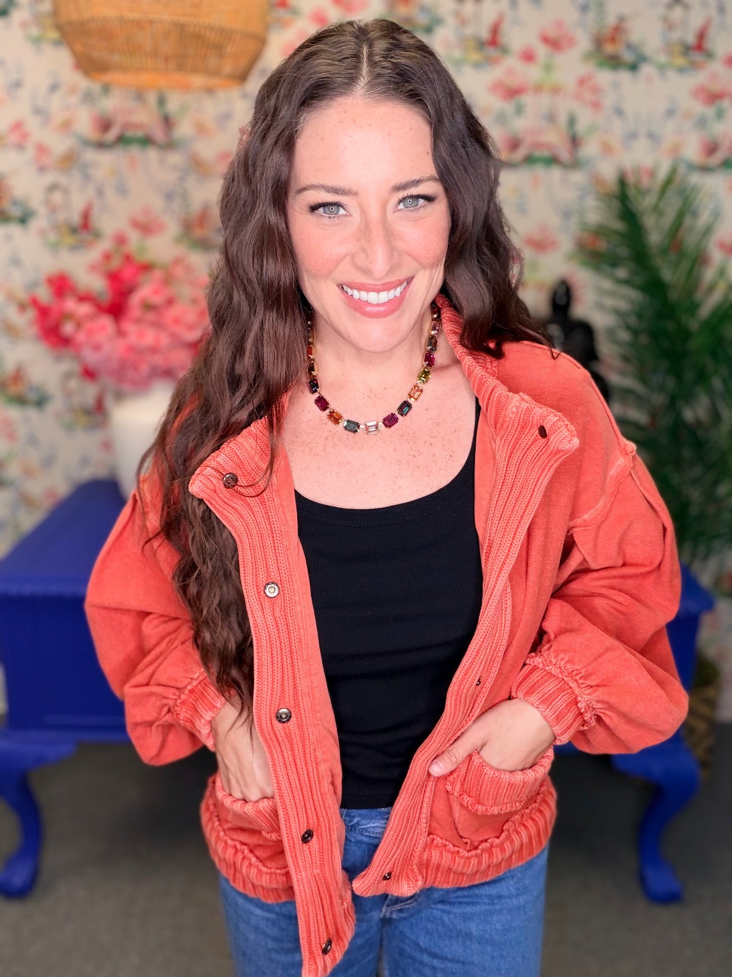 A smiling woman with long wavy brown hair stands indoors wearing a coral-orange corduroy jacket over a black tank top and blue jeans. The background features a floral wallpaper, a blue bench, and green plants.