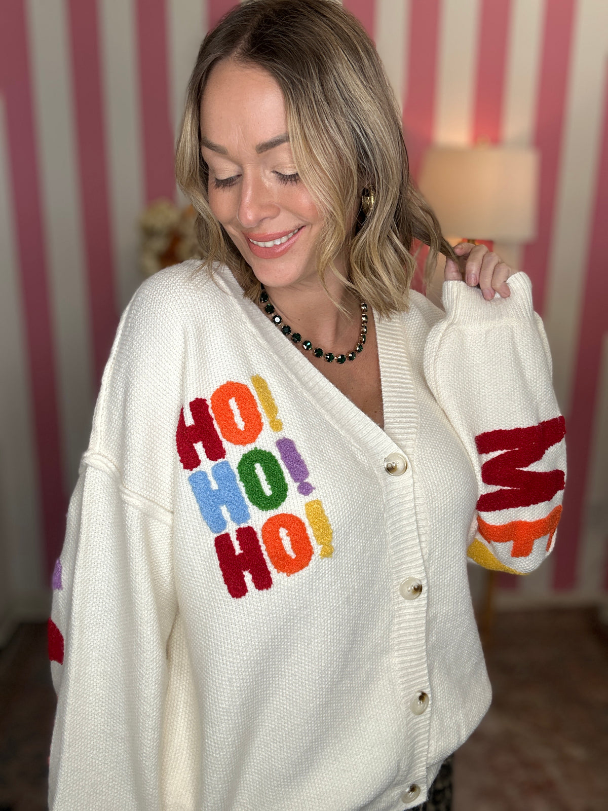A woman wearing a cream-colored knit cardigan sweater with colorful 'HO! HO! HO!' lettering on the front and 'Merry' on the sleeve, posed in front of a pink and white striped wall.