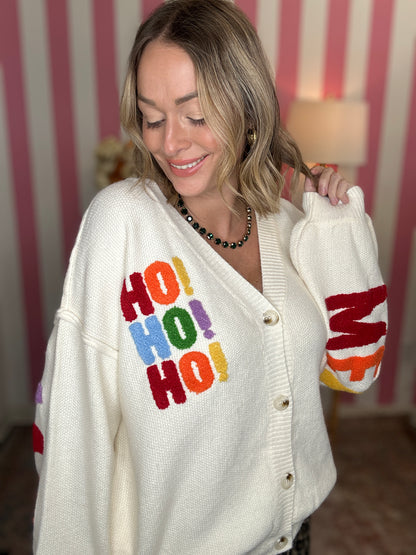 A woman wearing a cream-colored knit cardigan sweater with colorful 'HO! HO! HO!' lettering on the front and 'Merry' on the sleeve, posed in front of a pink and white striped wall.