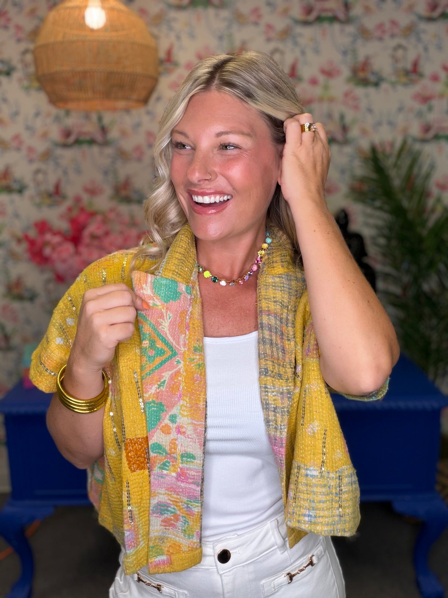 A smiling blonde woman wearing a yellow patterned knit cardigan over a white top, standing in front of floral wallpaper and a blue table.