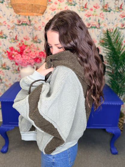 A woman wearing a light gray and dark brown patchwork sherpa fleece jacket, standing in front of a floral wallpaper background with a blue side table.