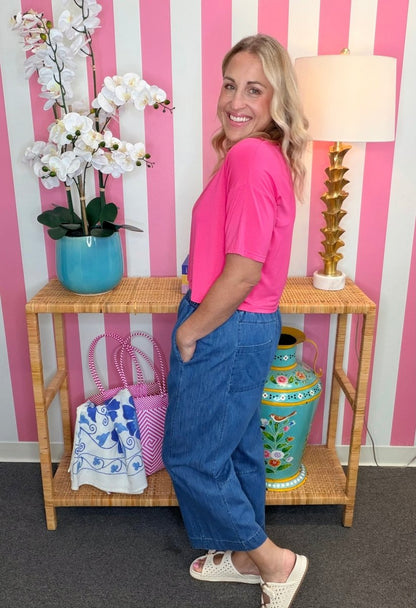 A woman modeling a bright pink short-sleeve crop top and wide-leg blue jeans, standing in front of a pink and white striped wall next to a bamboo console table with decorative items.