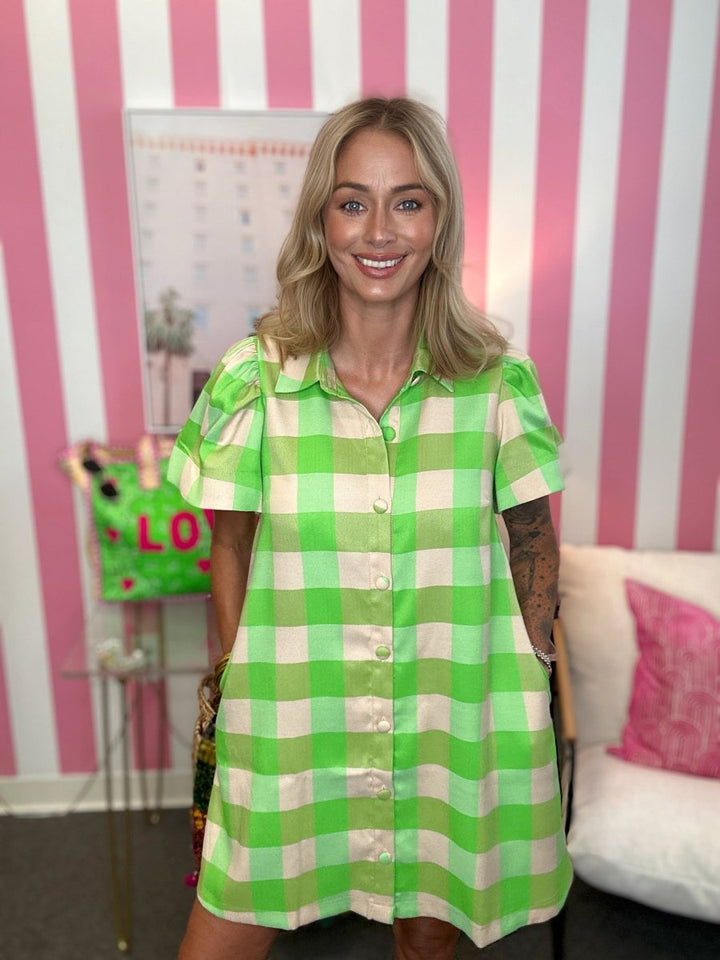 A woman wearing a bright green and white gingham short-sleeve button-up shirt dress, standing in front of a pink and white striped wall.