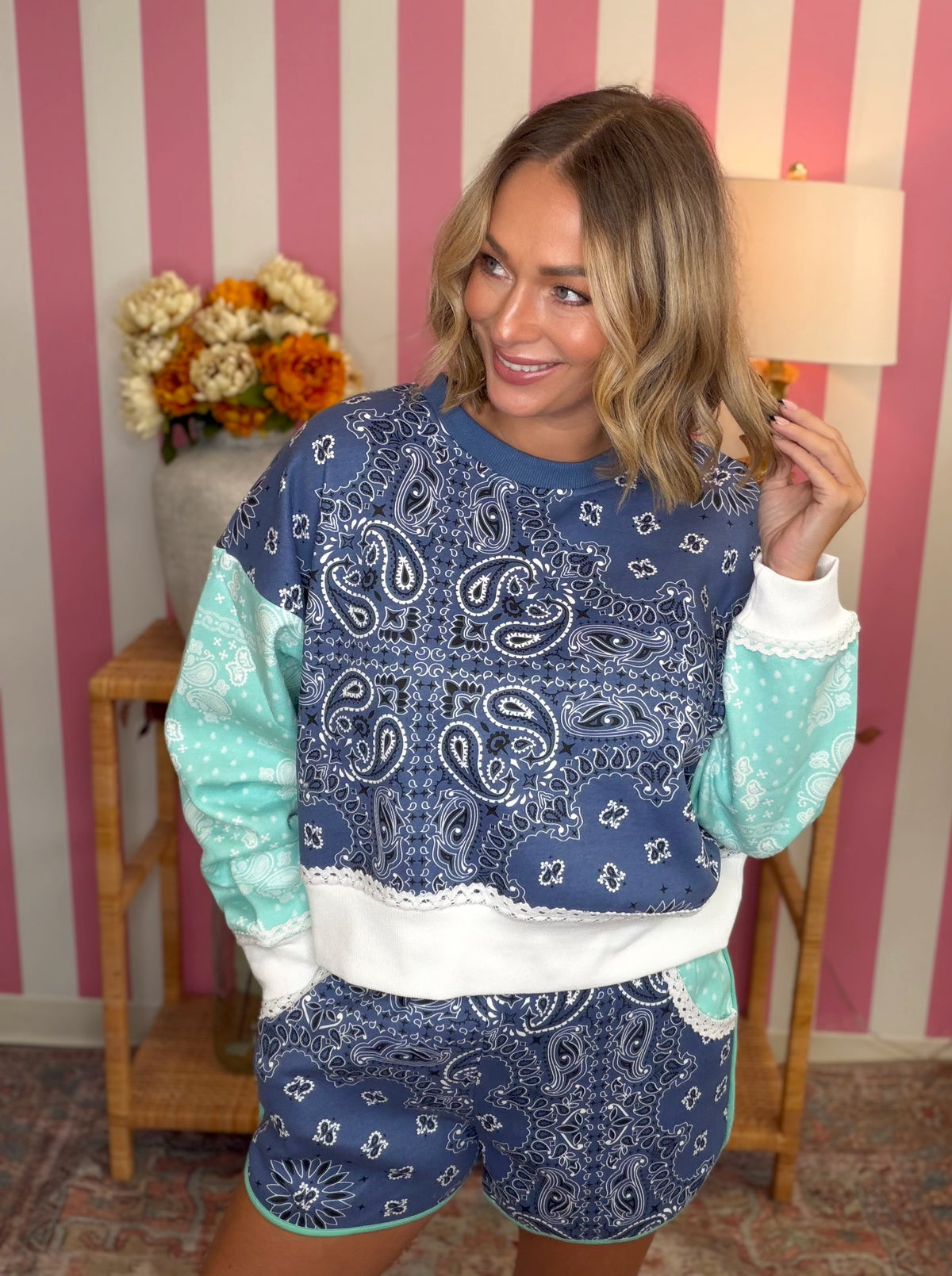 A woman modeling a matching blue paisley sweatshirt and shorts set with mint green sleeves and shorts, white lace trim, and a white waistband, set against a pink and white striped wall background.