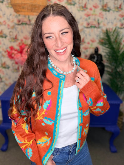 A woman smiling and wearing a bright orange knit cardigan with colorful abstract patterns in turquoise, pink, yellow, and blue, styled over a white top and blue jeans.
