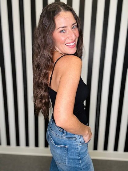 A woman with long wavy brown hair smiles while looking back over her shoulder, wearing a black spaghetti-strap camisole top and blue jeans, standing in front of a black and white vertically striped wall.