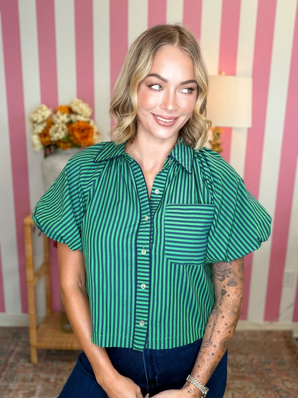 A woman wearing a green and dark green vertically striped short-sleeve button-up shirt with puff sleeves, paired with dark blue jeans, standing in front of a pink and white striped wall.