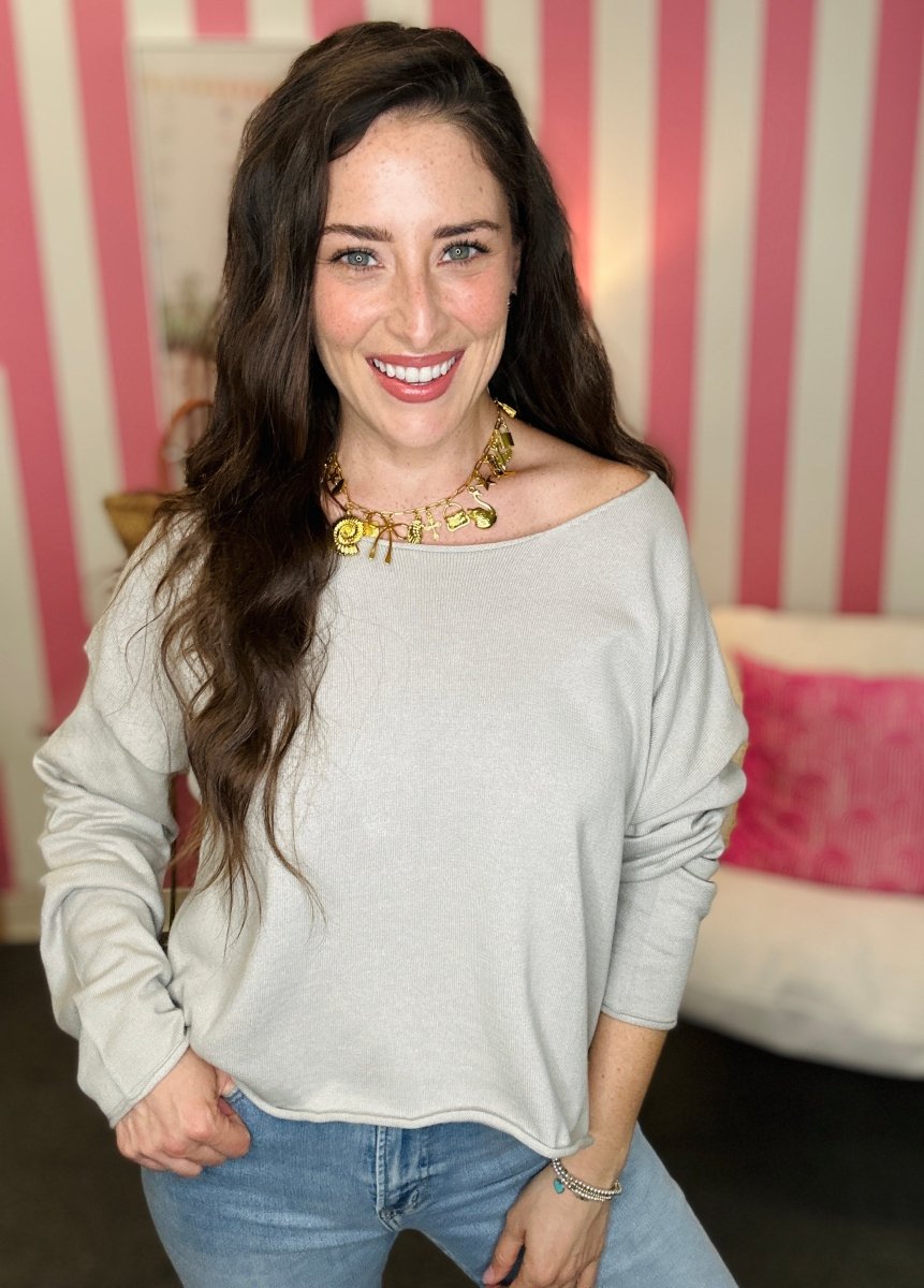 Woman wearing a light gray off-shoulder top and blue jeans with a striped wall in the background.