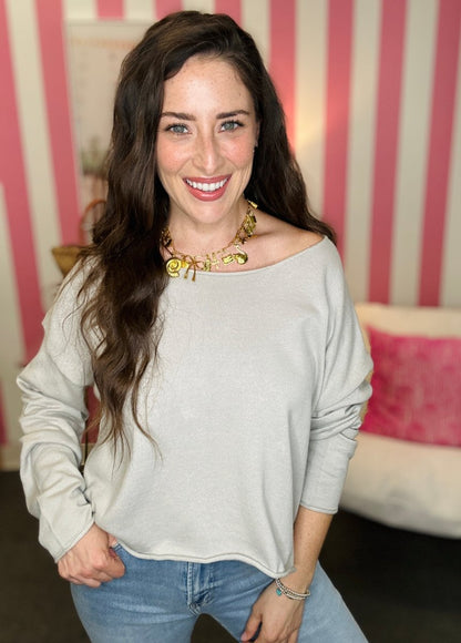 Woman wearing a light gray off-shoulder top and blue jeans with a striped wall in the background.