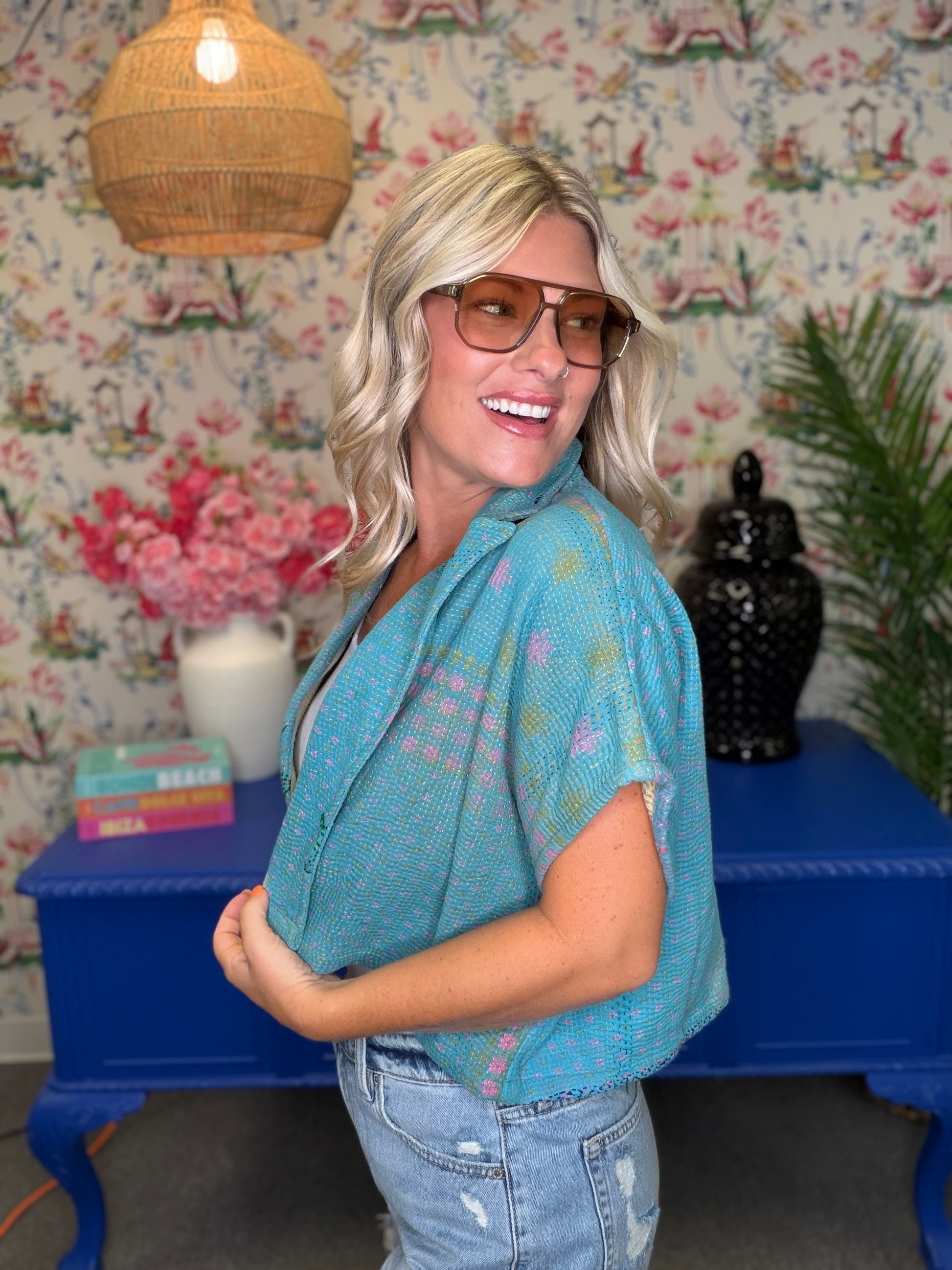 A woman with blonde hair and glasses wearing a light blue short-sleeve button-up shirt with colorful pastel star prints, paired with light-wash jeans, standing in front of a blue console table and floral wallpaper.