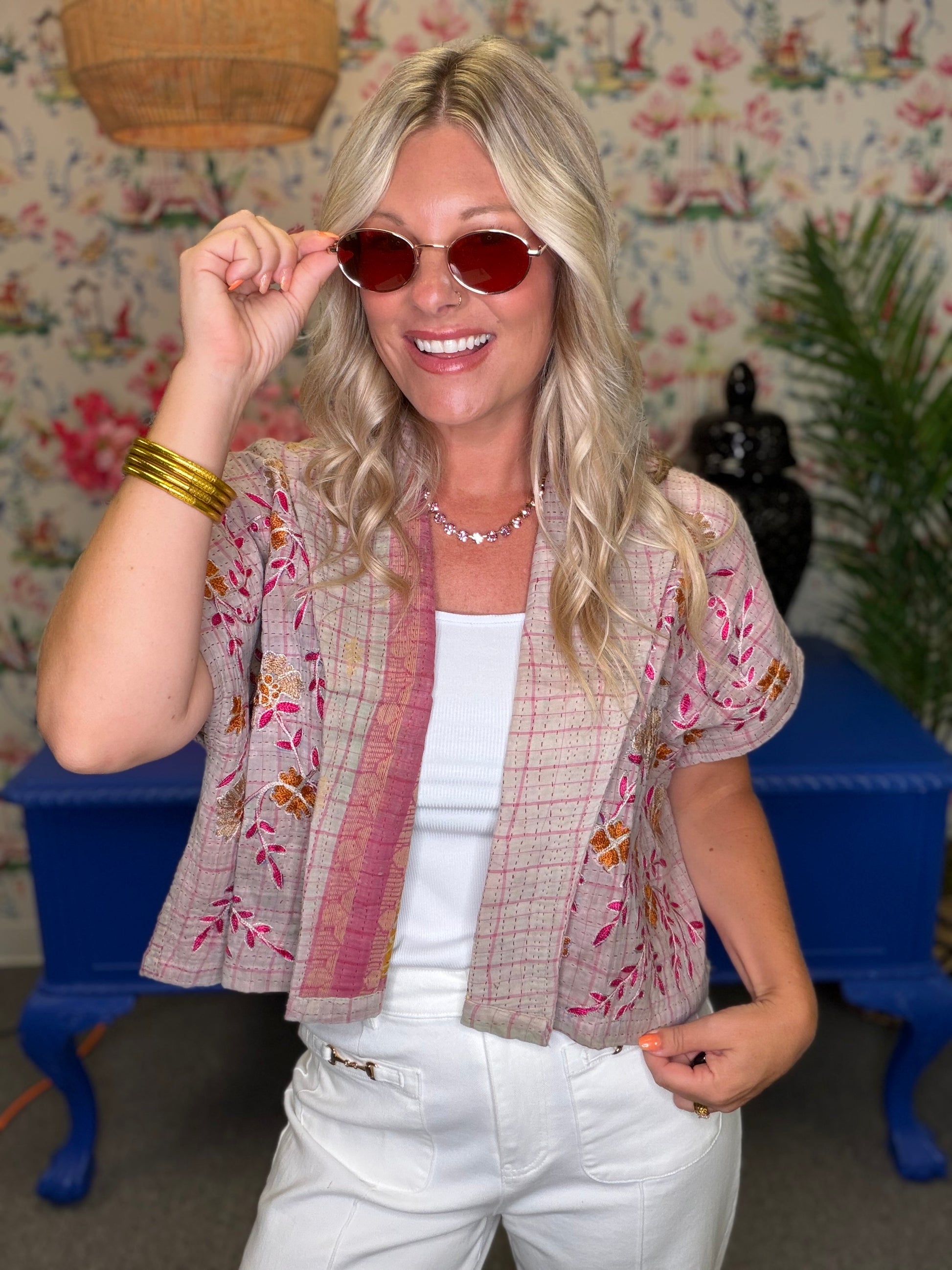 A woman wearing a beige and pink floral patchwork short-sleeve cardigan over a white tank top and white pants, posing in front of a floral wallpaper background with blue furniture.