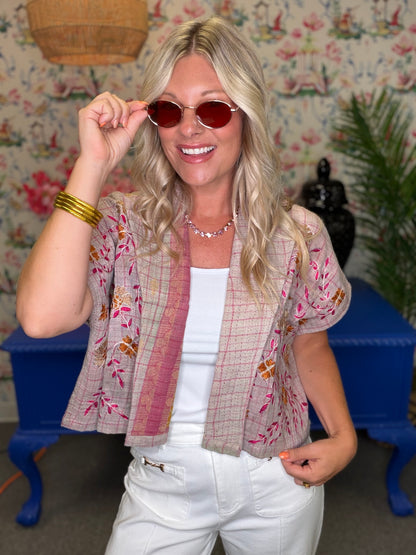A woman wearing a beige and pink floral patchwork short-sleeve cardigan over a white tank top and white pants, posing in front of a floral wallpaper background with blue furniture.