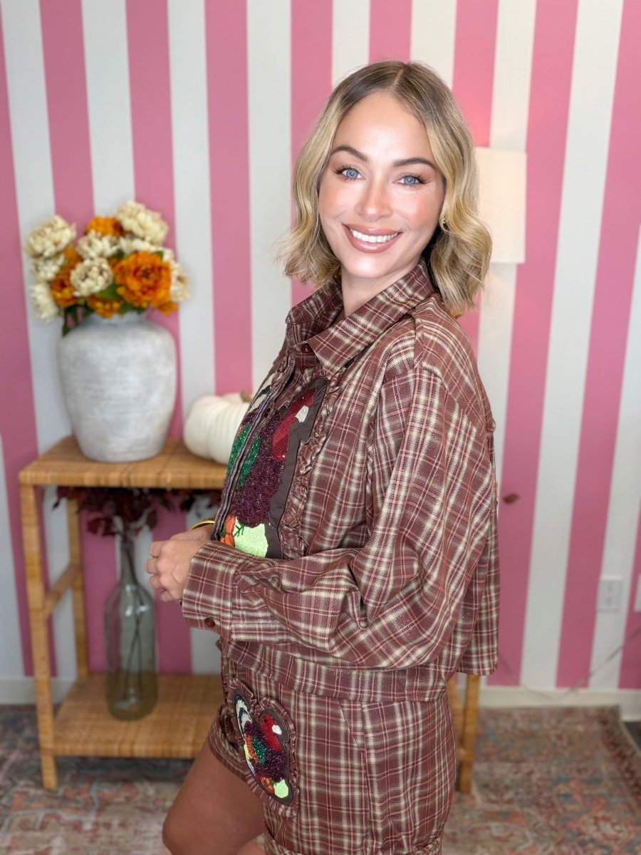 A woman with blonde wavy hair stands in front of a pink and white striped wall, wearing a brown and red plaid button-up shirt with a large embroidered patch on the pocket. She is standing next to a small wooden side table with a vase of flowers.