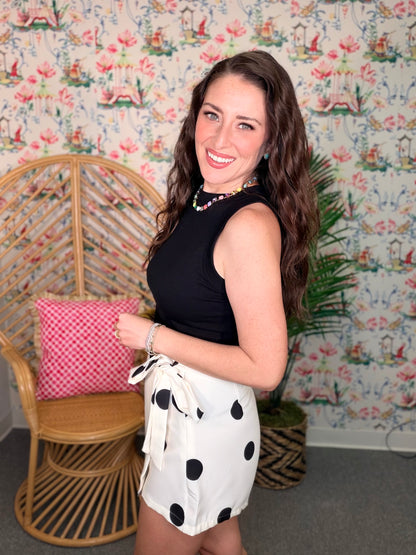 Woman in a black top and polka dot shorts standing in a room with floral wallpaper.
