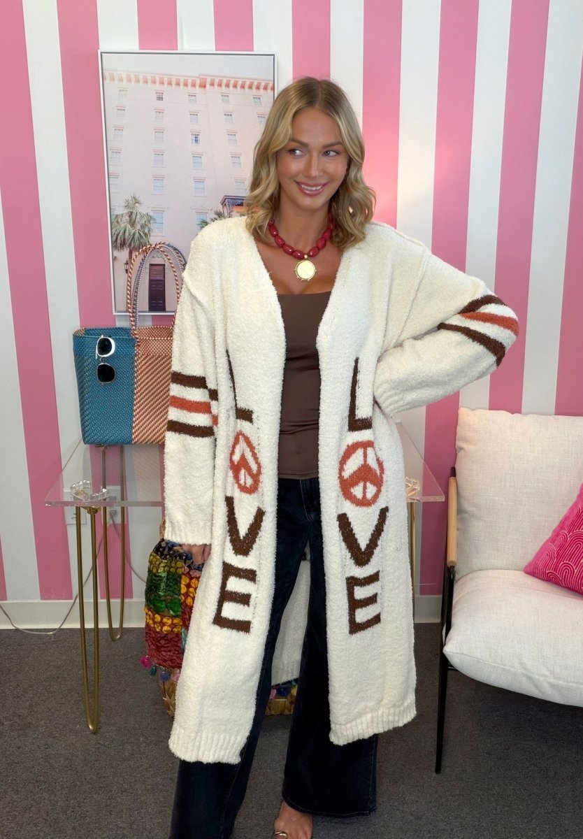 A woman models a long, cream-colored fuzzy cardigan with brown and red graphic details, including peace signs and the word 'LOVE' in brown letters. She is standing in a room with pink and white striped walls.