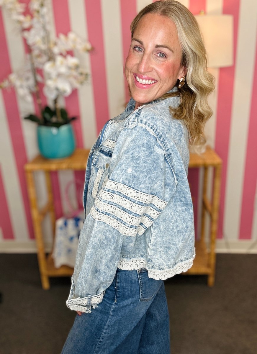 A woman wearing a light blue acid-wash cropped denim jacket with white lace trim on the sleeves and hem, paired with blue jeans, posing in front of a pink and white striped wall.