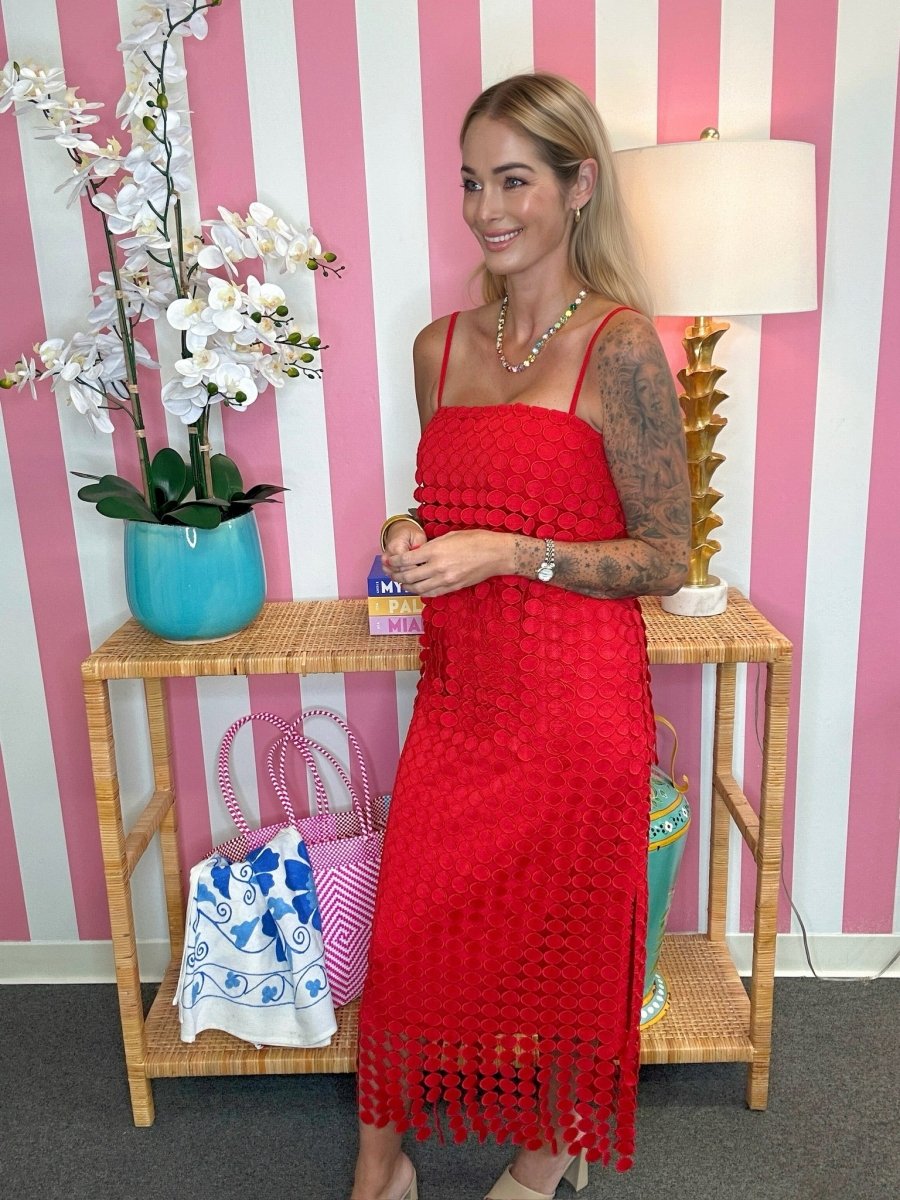 A woman wearing a bright red crocheted sleeveless dress with thin straps and a tassel fringe hem, standing indoors in front of pink and white striped wallpaper. She is posed next to a wooden side table with a turquoise orchid pot, a lamp, and patterned handbags.