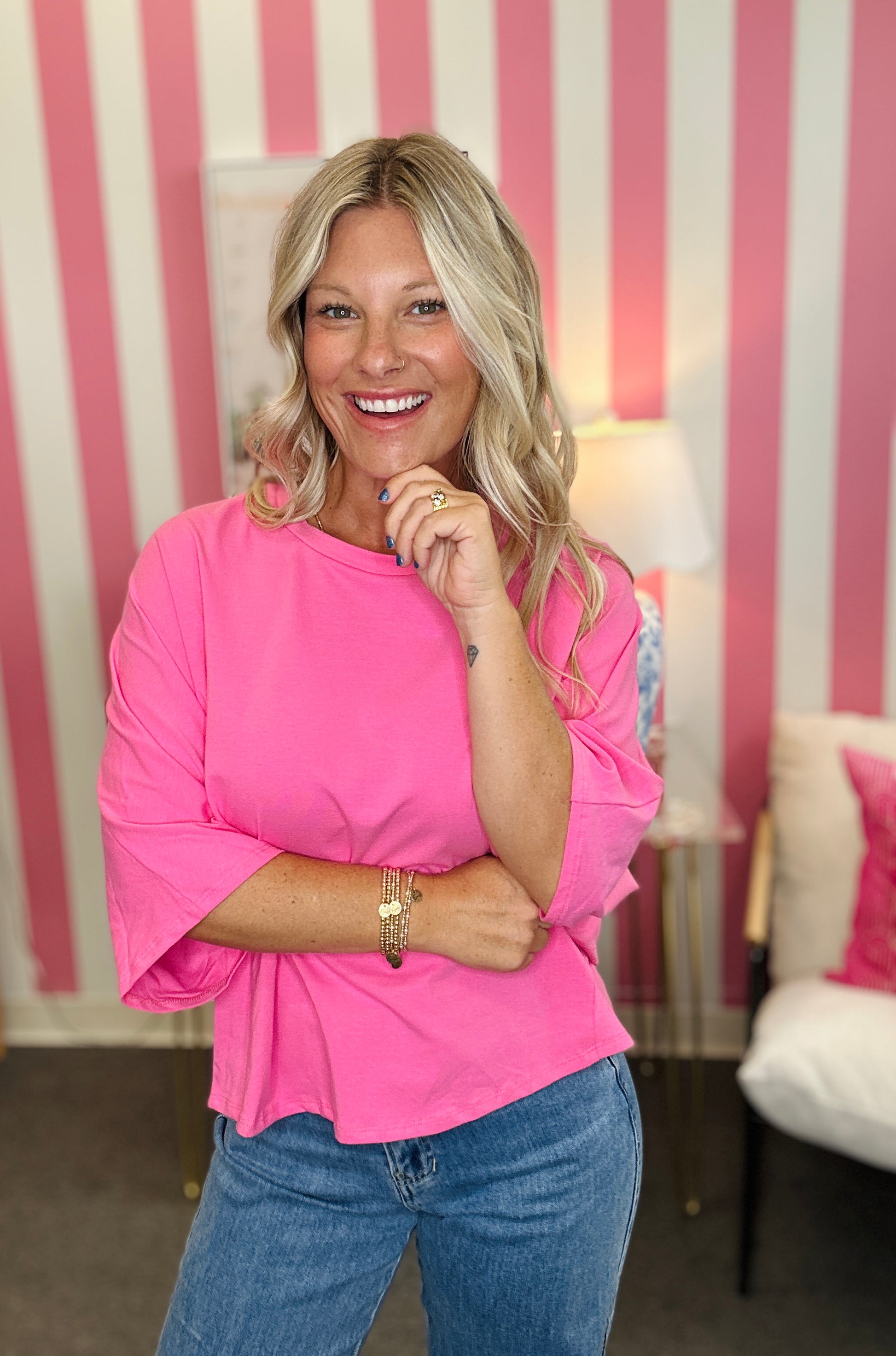 A smiling blonde woman wearing a bright pink three-quarter sleeve top and blue jeans, standing in front of a pink and white striped wall.