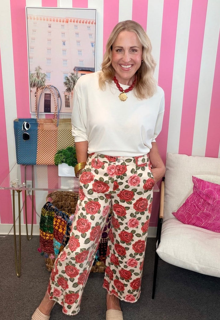 A woman stands in a room with pink and white striped walls, wearing a white long-sleeve top and cream wide-leg pants with a red and pink rose pattern. She is accessorized with a red beaded necklace and gold bracelets.