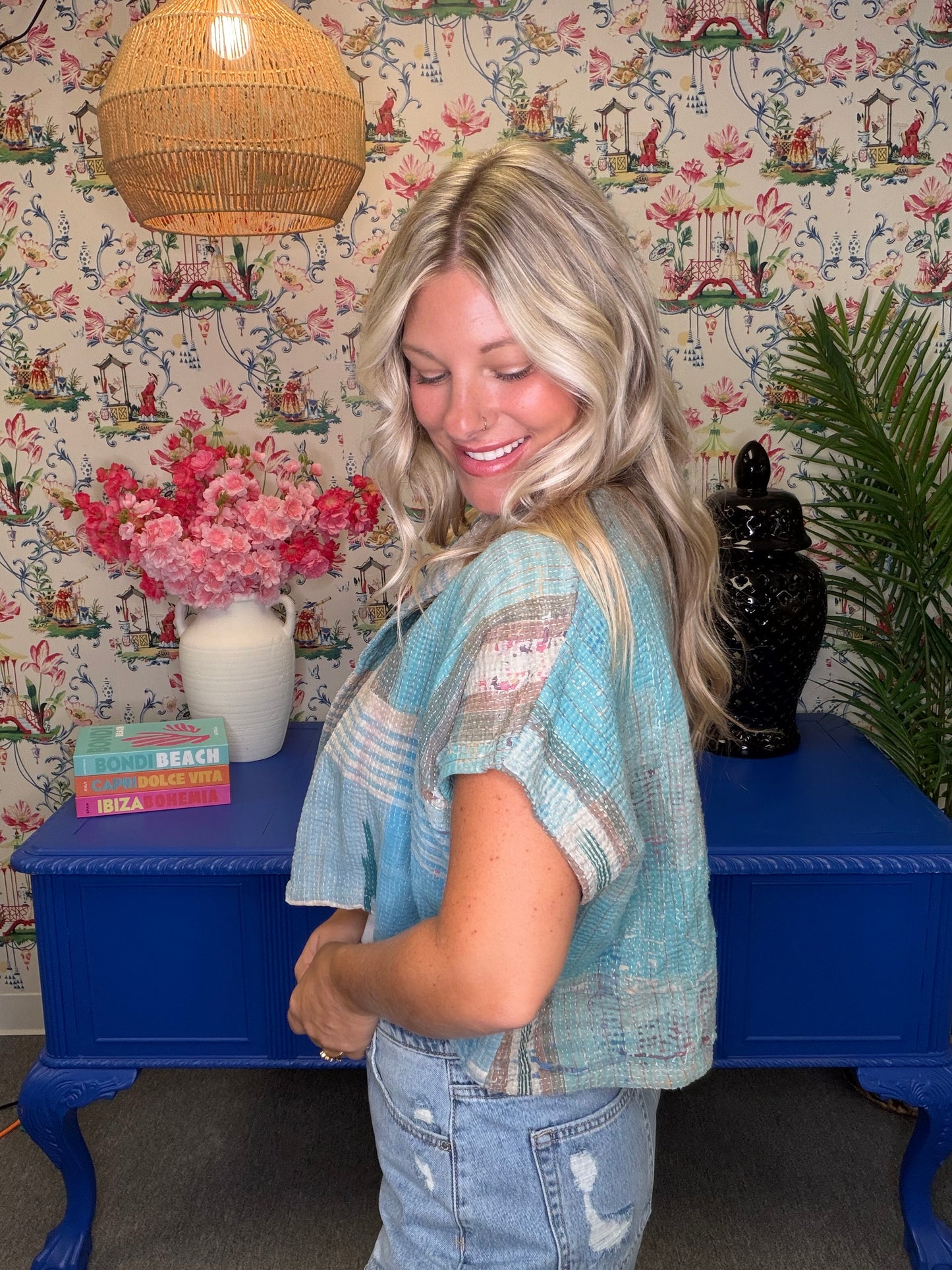 A woman with blonde hair wearing a light blue and beige plaid short-sleeve top and light-wash jeans, standing in front of a blue console table decorated with wallpaper, a vase of pink flowers, and a wicker pendant light.