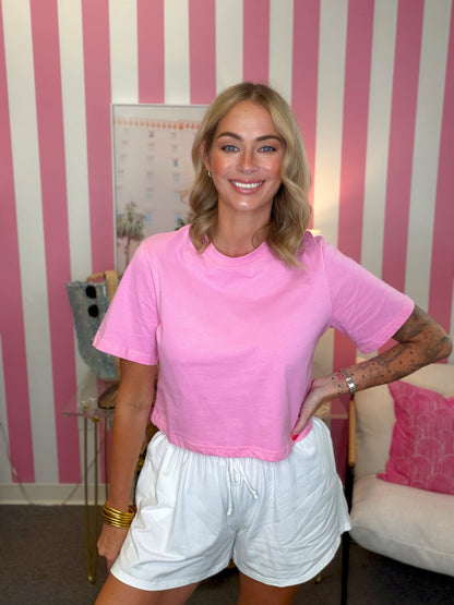 A woman posing in a bright pink short-sleeve crewneck t-shirt and white shorts, standing in front of a pink and white striped wall.