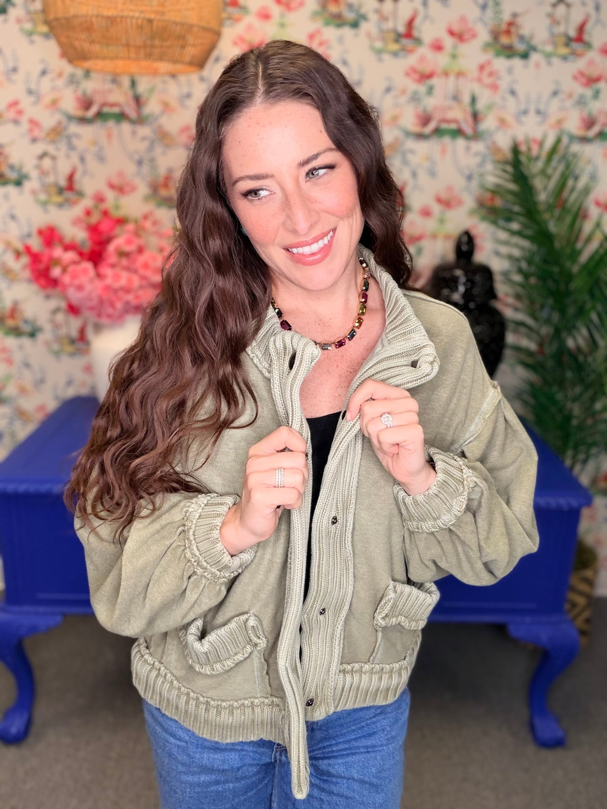 A woman with long wavy brown hair wearing an olive green button-up jacket with ruffled trim, posing in front of a floral wallpaper background with a blue bench.