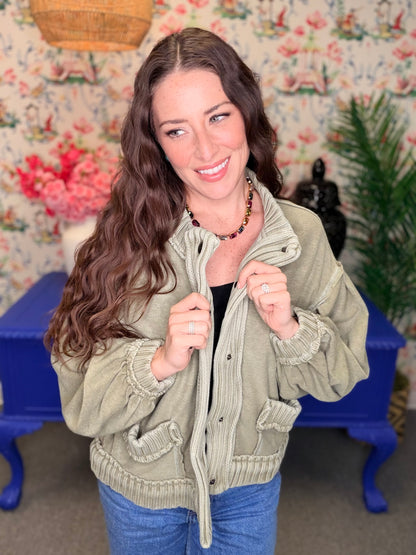 A woman with long wavy brown hair wearing an olive green button-up jacket with ruffled trim, posing in front of a floral wallpaper background with a blue bench.