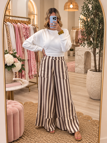 A woman takes a mirror selfie in a boutique, wearing a white long-sleeve crop top and brown-and-white vertically striped wide-leg pants with large patch pockets.