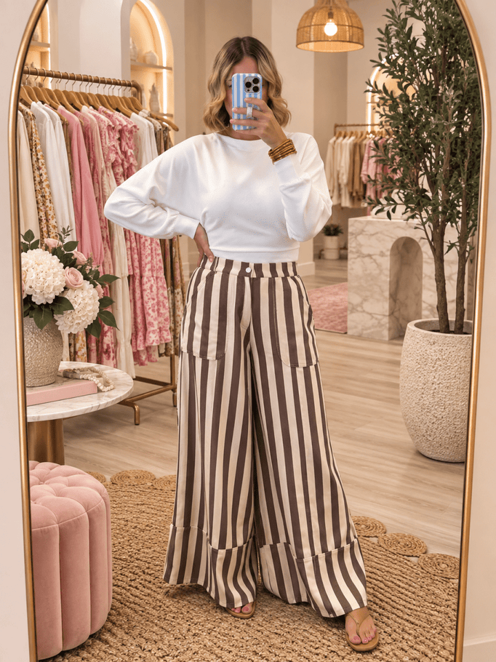 A woman takes a mirror selfie in a boutique, wearing a white long-sleeve crop top and brown-and-white vertically striped wide-leg pants with large patch pockets.