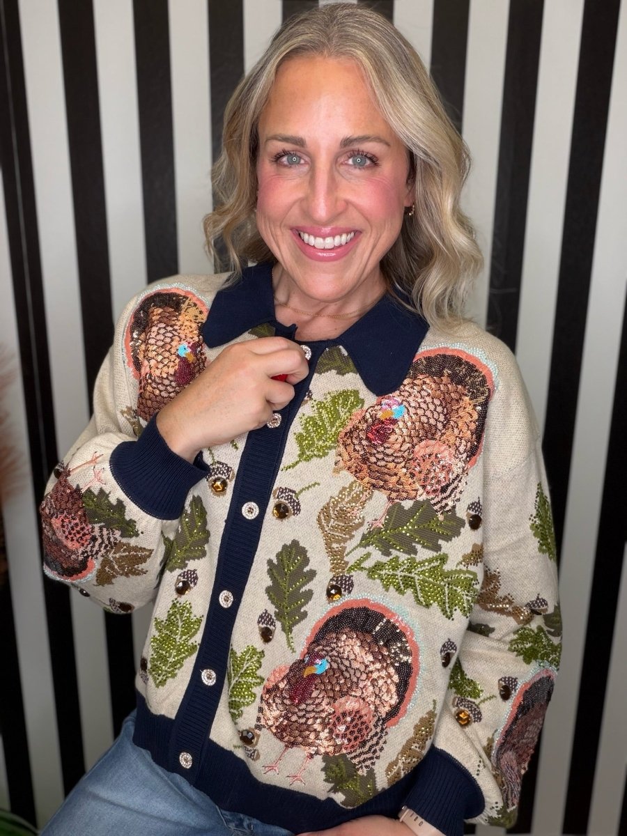 A smiling blonde woman wearing a beige knit button-up cardigan with a navy collar, cuffs, and button placket, decorated with large brown and green turkey and acorn motifs, posed against a black and white vertically striped background.