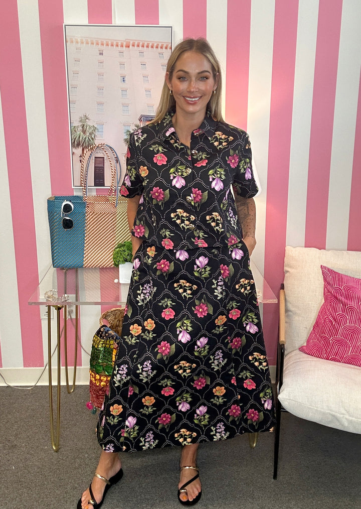 A woman wearing a black midi dress with a colorful floral pattern stands in front of a pink and white striped wall. She is wearing black sandals, and the background features a glass table with handbags, a framed print, and a white sofa with a pink pillow.