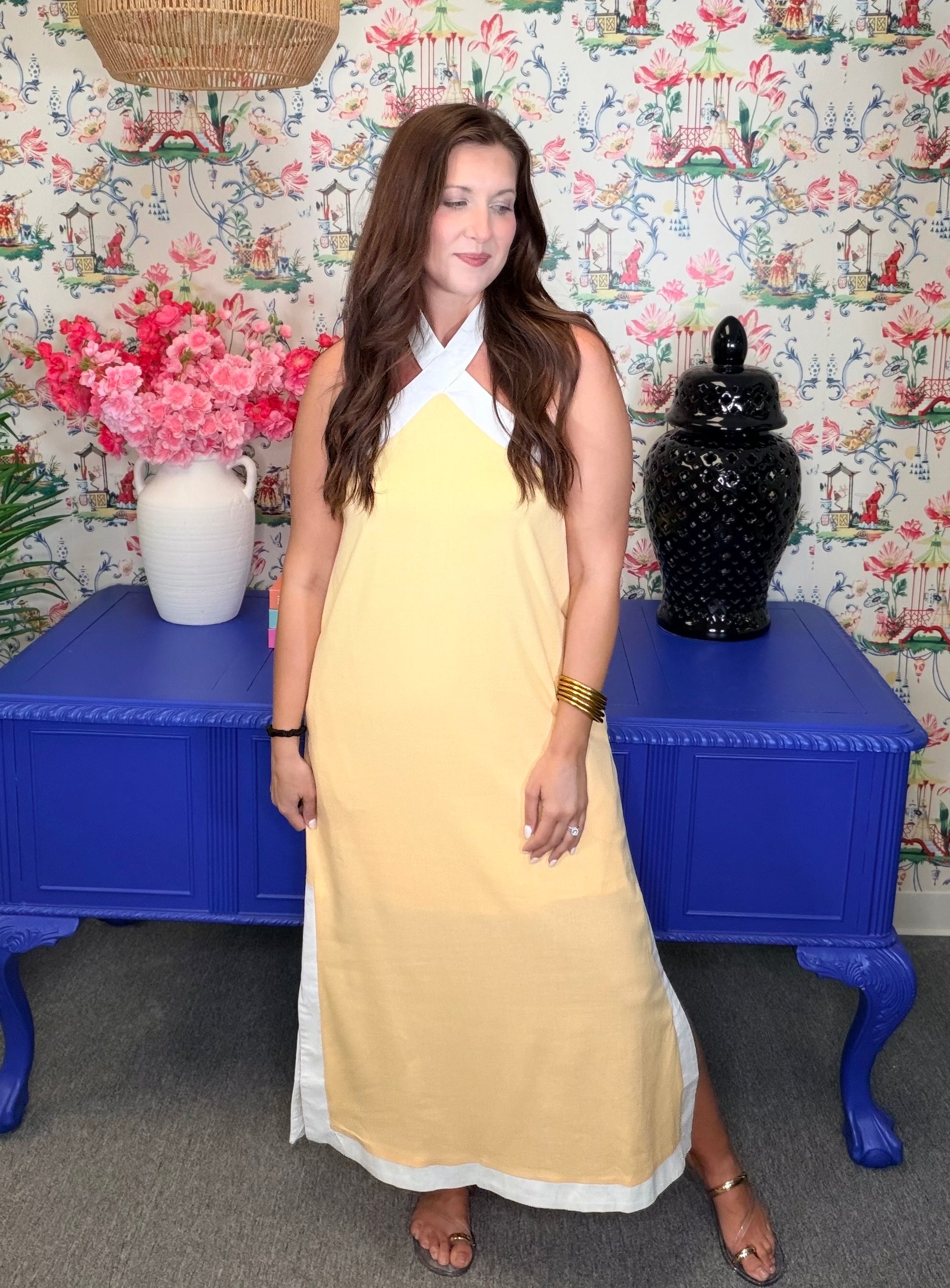 Woman in a yellow dress standing in front of a colorful floral-patterned wall with decorative vases and flowers.