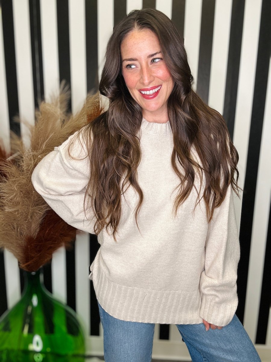 A woman with long wavy brown hair wearing a cream-colored knit sweater and blue jeans, standing in front of a black and white striped background.
