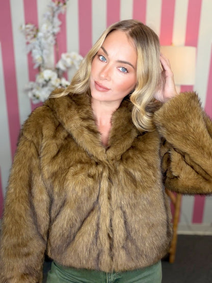 A woman wearing a brown faux fur jacket with a wide collar, posed in front of a pink and white striped wall.