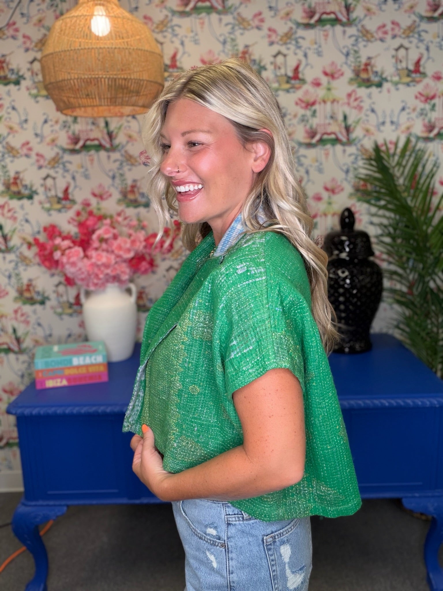 A woman wearing a bright green textured short-sleeve cropped jacket over a light blue collared shirt and light wash jeans, standing in front of a blue console table and floral wallpaper.