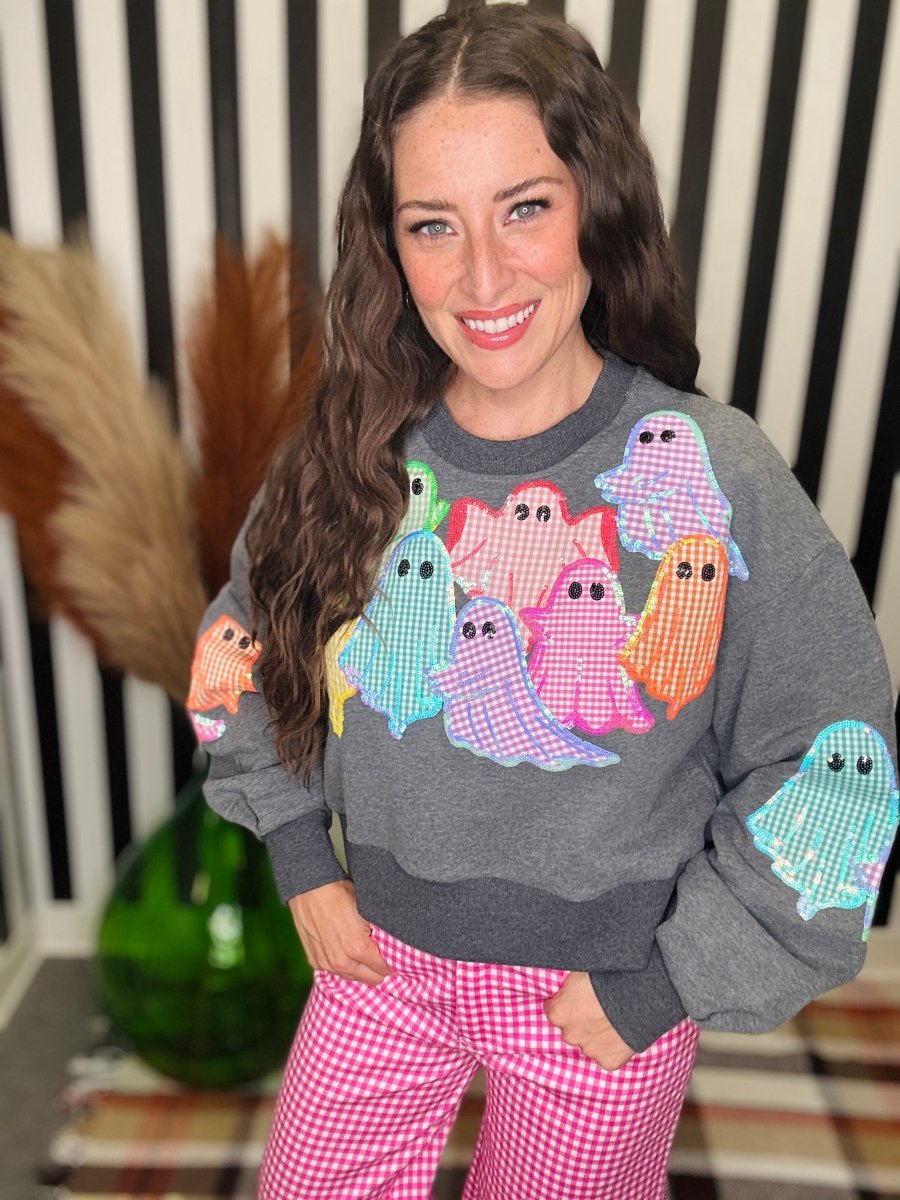 A woman wearing a gray crewneck sweatshirt with colorful gingham-patterned ghost appliqués, paired with pink and white gingham pants, standing in front of a black and white striped background.