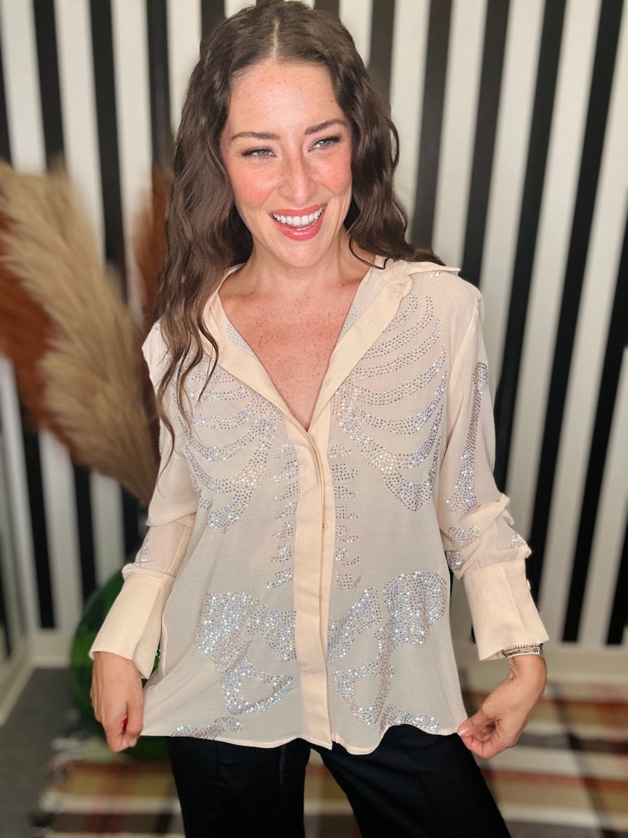 A woman wearing a sheer beige button-up blouse with silver sequin embellishments, paired with black pants, standing in front of a black and white striped background.