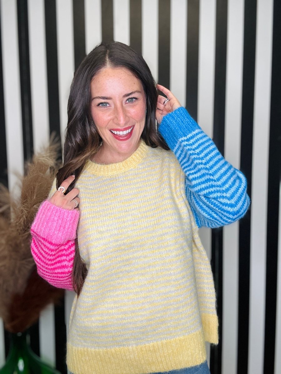 A woman modeling a chunky knit sweater with a yellow body and contrasting sleeves. One sleeve is bright pink with horizontal stripes, and the other is blue with horizontal stripes.