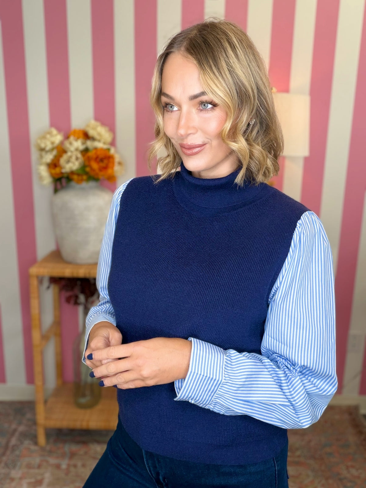 A woman wearing a navy blue ribbed turtleneck sweater vest over a light blue and white striped long-sleeve shirt, standing in front of a pink and white striped wall.