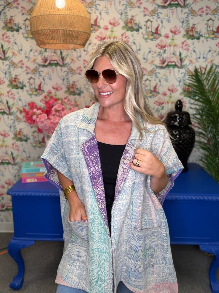 A woman wearing a light blue and lavender patterned open-front cardigan over a black top, standing in front of a floral wallpaper background with a blue cabinet.