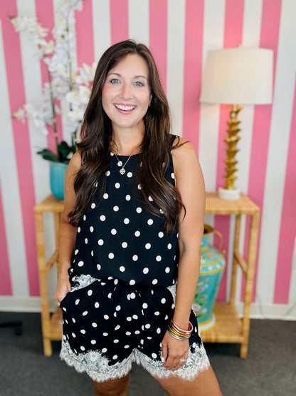 Woman wearing a black polka dot top and white lace shorts against a pink and white striped wall.