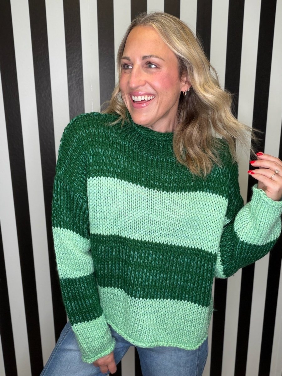 A woman wearing a chunky knit sweater with horizontal stripes in dark forest green and light mint green, paired with blue jeans, standing in front of a black and white striped wall.