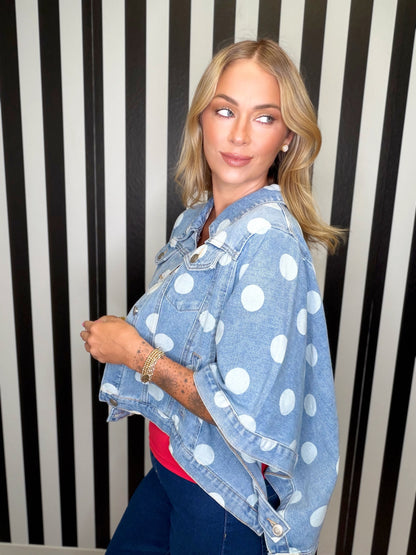 A woman wearing a light blue denim jacket with large white polka dots, styled over a red top and dark blue jeans, posing in front of a black and white striped wall.