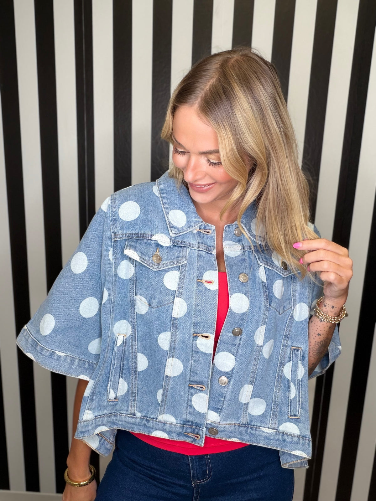 A woman wearing a light blue cropped denim jacket with large white polka dots, layered over a bright pink top and dark blue jeans, standing against a black and white striped background.