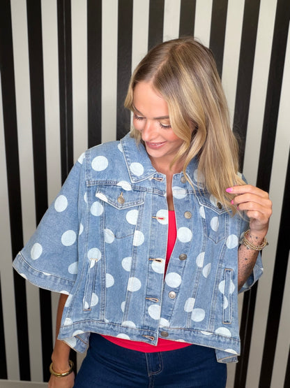 A woman wearing a light blue cropped denim jacket with large white polka dots, layered over a bright pink top and dark blue jeans, standing against a black and white striped background.