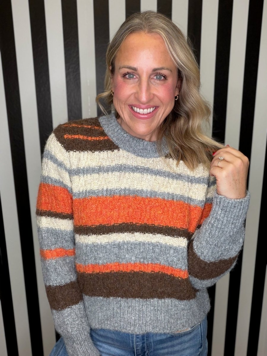 A smiling woman with blonde hair wearing a fuzzy knit sweater with horizontal stripes in gray, brown, orange, and cream, standing in front of a black and white striped wall.