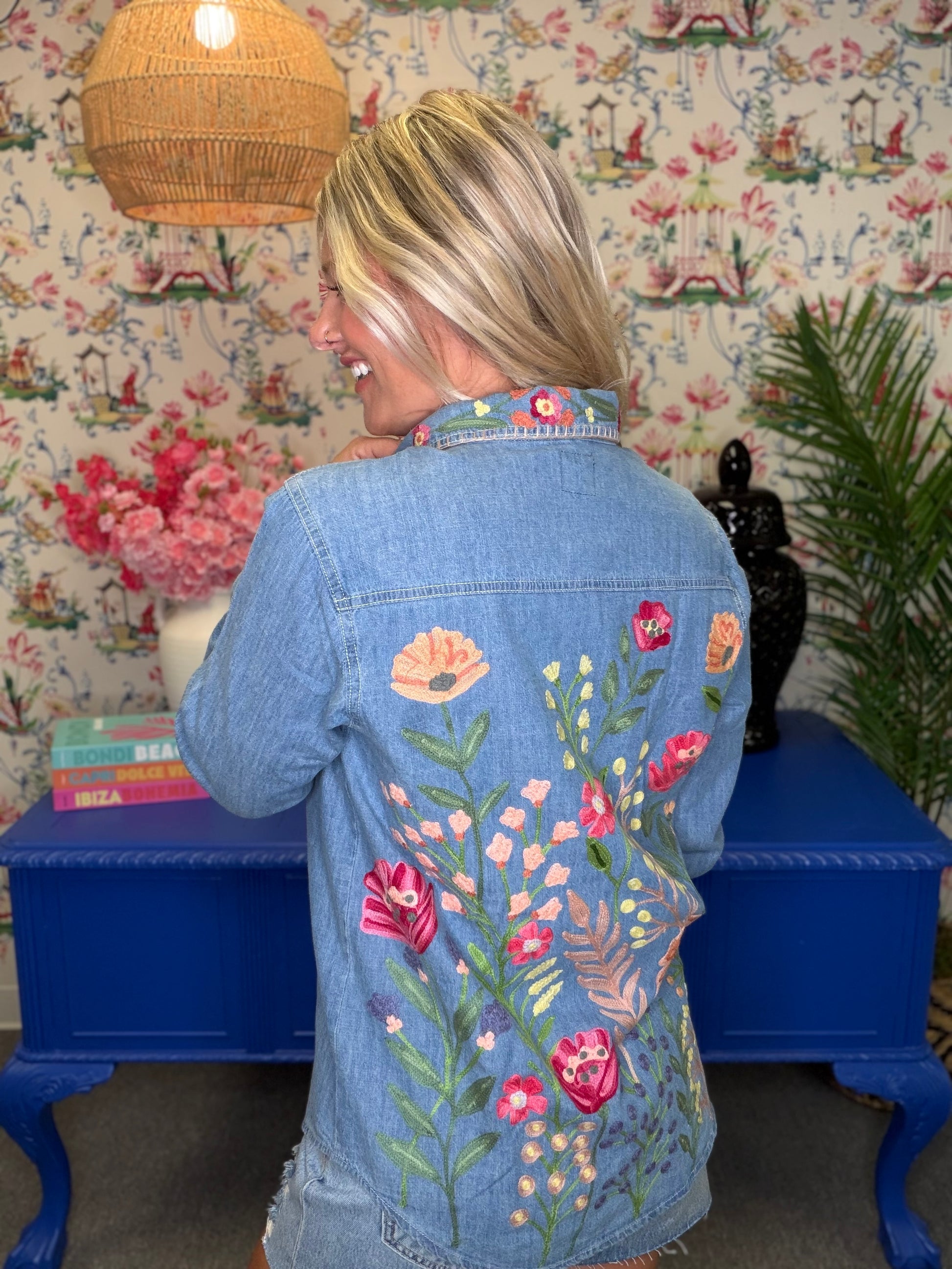 A woman viewed from behind wearing a light blue denim jacket with colorful floral embroidery on the back, standing in front of a blue table and floral wallpaper.