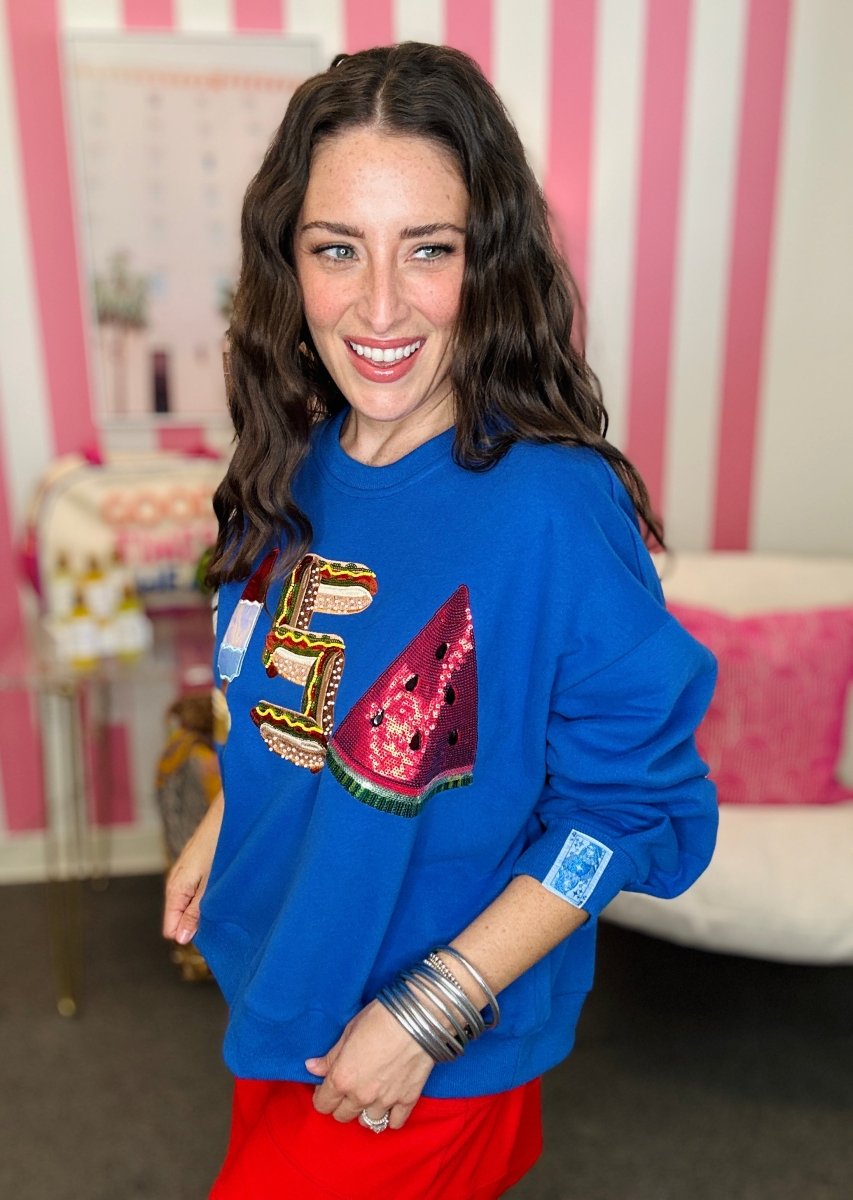 Woman wearing a blue sweatshirt with colorful designs in a room with pink and white striped wallpaper.